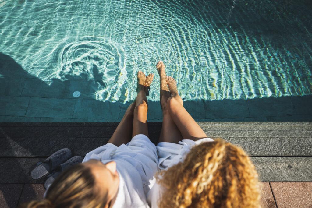 Two girlfriends dangle their feet in the turquoise pool water of the Hotel Klosterbräu. They wear white bathrobes