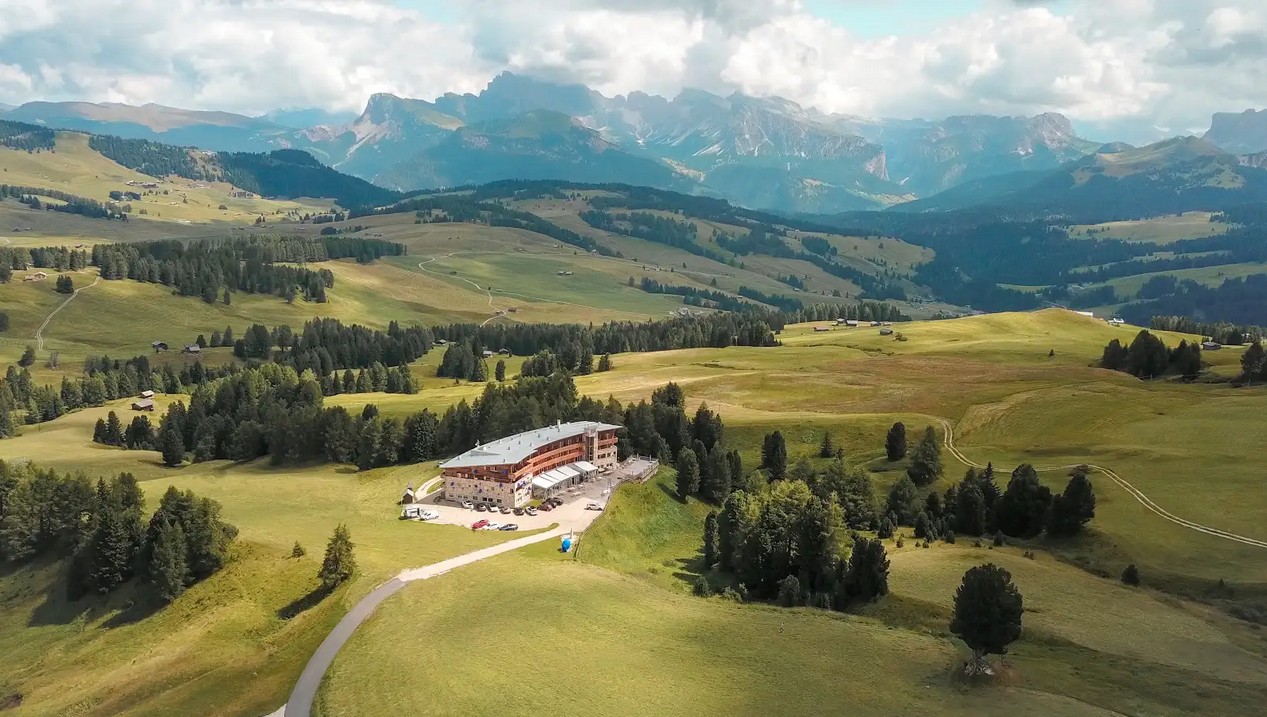 Eine ruhige Berglandschaft mit dem Paradiso Pure.Living Vegan Hotel, umgeben von weitläufigen grünen Feldern und vereinzelten Wäldern. Sanfte Hügel und ferne Berge unter einem teilweise bewölkten Himmel schaffen eine friedliche Atmosphäre.