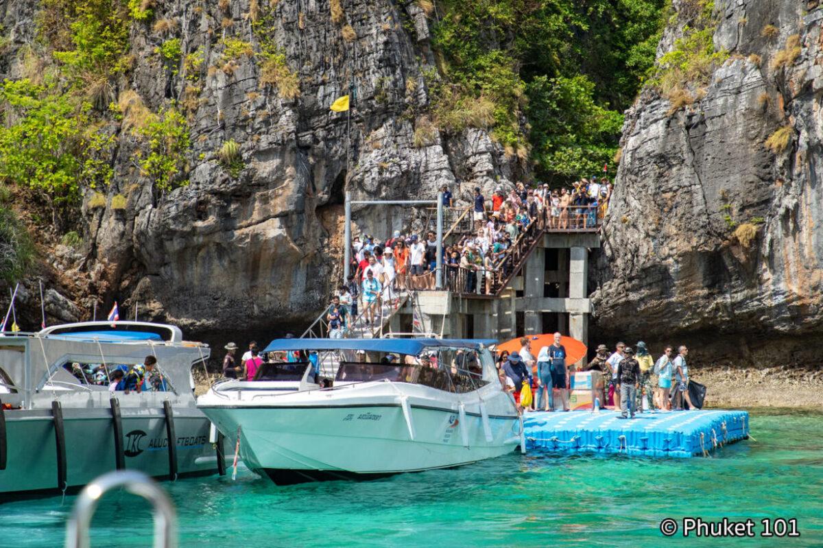 Tourists are crowding onto a staircase, and in the foreground are boats in Maya Bay, Thailand, in 2026. Maya Bay became famous as the filming location for “The Beach.”