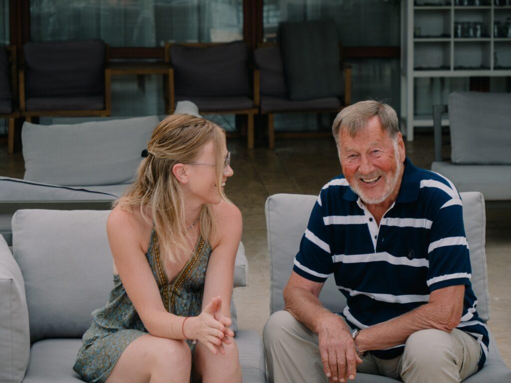 A young woman and an older man are sitting on an outdoor sofa, smiling and chatting. The setting is relaxed, with furniture in neutral tones.