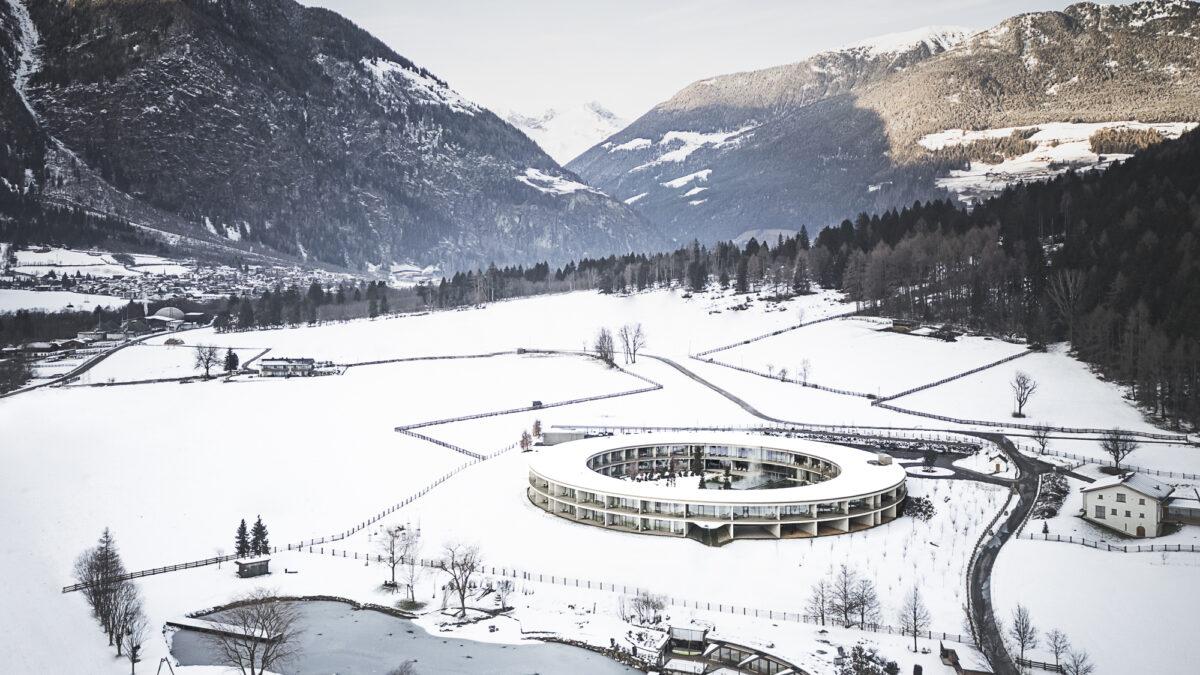 Aerial view of a modern, circular building surrounded by snow-covered fields and mountains, with a peaceful winter atmosphere and clear blue skies.