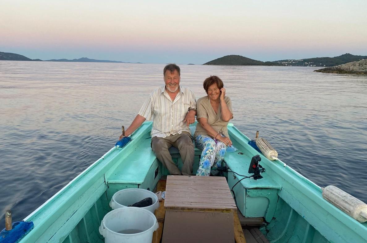 A couple sits smiling in a turquoise boat on calm waters as the sun sets. The horizon is dotted with distant islands, creating a peaceful, serene scene.