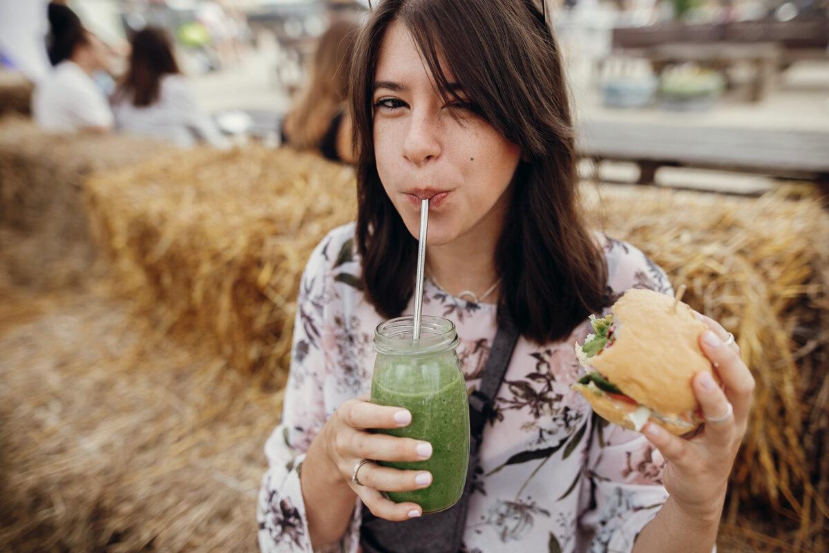 Stylish hipster girl holding a delicious vegan burger and drinking a smoothie from a glass jar at a street food festival. Happy boho woman tasting a burger with a healthy drink on a summer street.
