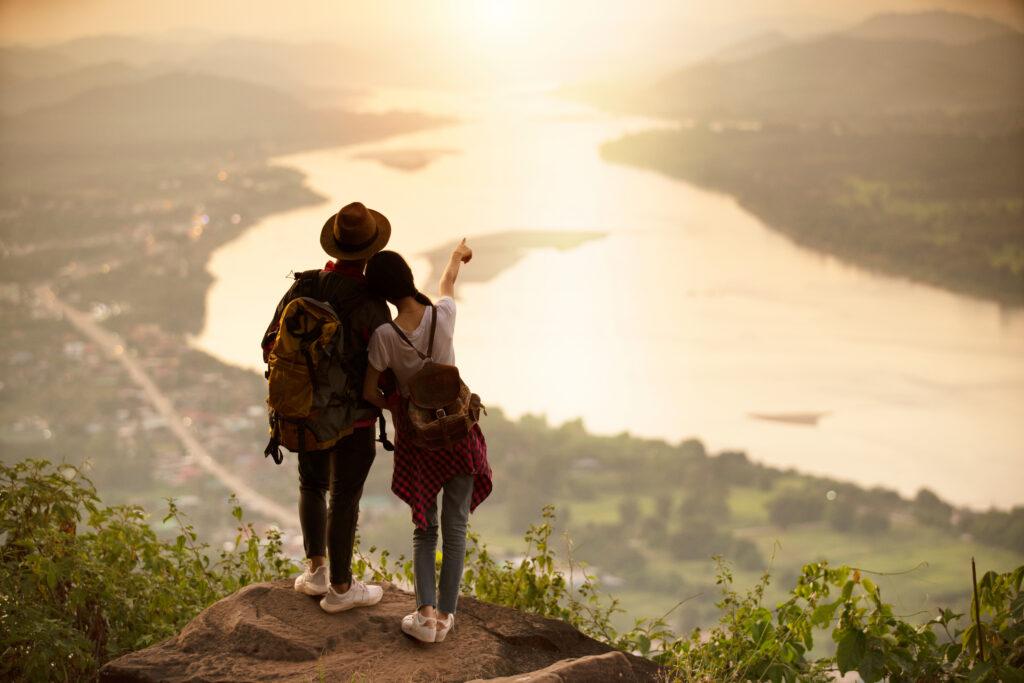 Two travelers stand on a rocky outcrop and look into the sunset, which colors the river below them gold.